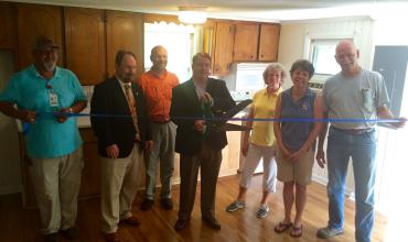 group of people around a ribbon getting ready to cut it inside a home