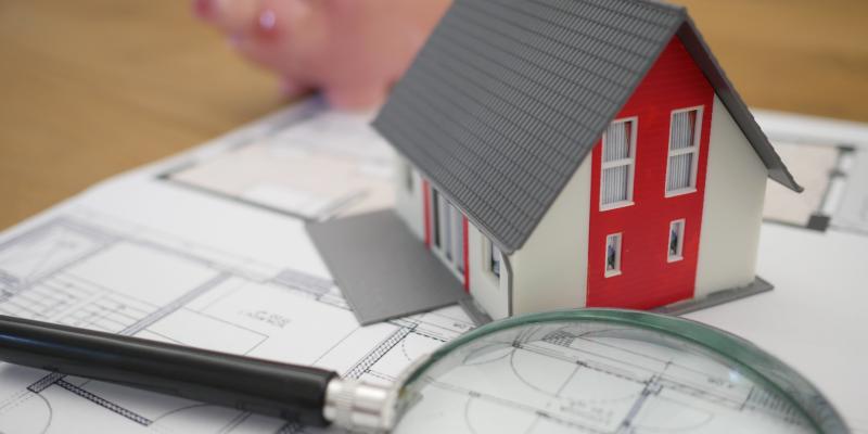 A small red house sitting on blueprints next to a microscope and a piggy bank